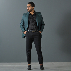 Hombre posando con un blazer premium slim fit verde inglés, camisa negra y pantalones oscuros, en un fondo gris.