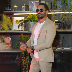 Hombre vestido con un traje beige y gafas de sol, posando en un ambiente decorado con plantas y estantes con libros.