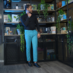 Hombre de pie en una habitación moderna, vistiendo un suéter negro y pantalones azules claros, rodeado de plantas y estantes con productos.