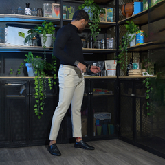 Hombre de pie en una habitación moderna, vistiendo un suéter negro y pantalones claros, interactuando con estantes llenos de plantas y objetos decorativos.