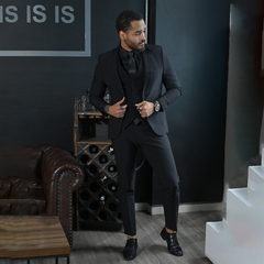 Hombre elegante vestido con traje negro, posando en un ambiente moderno con un sofá de cuero marrón y una estantería de vino.