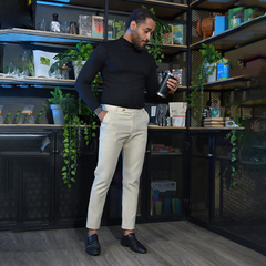 Hombre de pie sosteniendo un termo negro, vestido con un suéter negro y pantalones claros, en un entorno moderno con plantas y estanterías.
