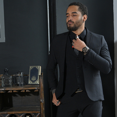 Hombre vestido con traje negro, ajustándose la corbata y mostrando anillos y reloj, en un entorno elegante.