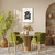 Quadro decorativo Matisse com fundo claro e desenho de flor preta, exposto em uma sala de estar com mesa redonda e cadeiras verdes.