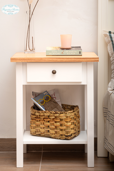 Mesa de luz Felipe con superficie de madera y estructura blanca, decorada con un jarrón, una vela y libros, junto a una cesta de mimbre con revistas.