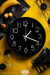 Reloj de pared negro con números blancos sobre fondo amarillo, acompañado de un plátano, frutas y utensilios de cocina.
