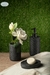 Set de baño negro con dispensador, jarrón con flores y pastilla de jabón sobre una superficie de madera.