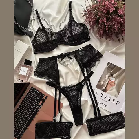 Conjunto de lingerie preto com detalhes em renda, incluindo sutiã, calcinha e liga, dispostos sobre uma superfície clara ao lado de um laptop e produtos de beleza.