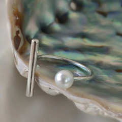 Anillo de plata delicado con diseño abierto, decorado con una perla blanca y una barra vertical, sobre concha iridiscente.