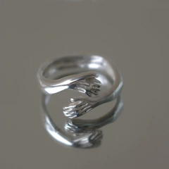 Anillo Hug de plata con diseño de manos entrelazadas, reflejándose en una superficie. Parte de la Colección Silhouette.