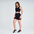 Modelo vestindo top esportivo feminino preto da Marcio May, combinado com shorts pretos, posando em um estúdio.
