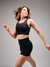 Modelo vestindo top esportivo feminino preto e shorts, posando com um sorriso em um fundo neutro.