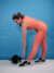 Mulher se exercitando com calça legging laranja e tênis preto, inclinando-se para pegar um haltere no chão.