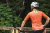 Homem usando uma camisa de ciclismo laranja, observando a paisagem com uma bicicleta ao seu lado.