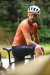 Homem sentado em uma bicicleta, vestindo uma camisa de ciclismo laranja com detalhes verdes e bermuda preta, em um ambiente ao ar livre.