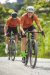 Ciclistas usando camisas de ciclismo em um caminho de terra, com a ciclista sorrindo e pedalando à frente.