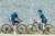 Casal de ciclistas em roupas de ciclismo azul, parados em bicicletas em uma estrada, com uma parede texturizada ao fundo.