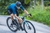 Homem em uma bicicleta de estrada, vestindo uma camisa de ciclismo azul, pedalando em uma estrada com vegetação ao fundo.