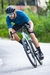 Homem pedalando uma bicicleta em uma estrada, vestindo a camisa de ciclismo Márcio May Pro Petroleo na cor azul.