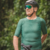 Homem sorrindo usando uma camisa de ciclismo verde, com capacete e óculos escuros, posando ao lado de uma bicicleta em um ambiente natural.