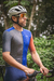 Homem usando camisa de ciclismo azul e cinza, com detalhe em laranja, segurando guidão de bicicleta em ambiente natural.
