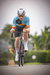 Atleta em bicicleta com camisa de ciclismo da Bélgica, pedalando em estrada com vegetação ao fundo.
