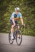 Homem montado em uma bicicleta, usando uma camisa de ciclismo azul com detalhes em vermelho e amarelo, pedalando em uma estrada.