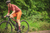 Homem montado em uma bicicleta de gravel, usando bretelle masculino na cor laranja e camiseta de manga curta, pedalando em uma trilha verde.