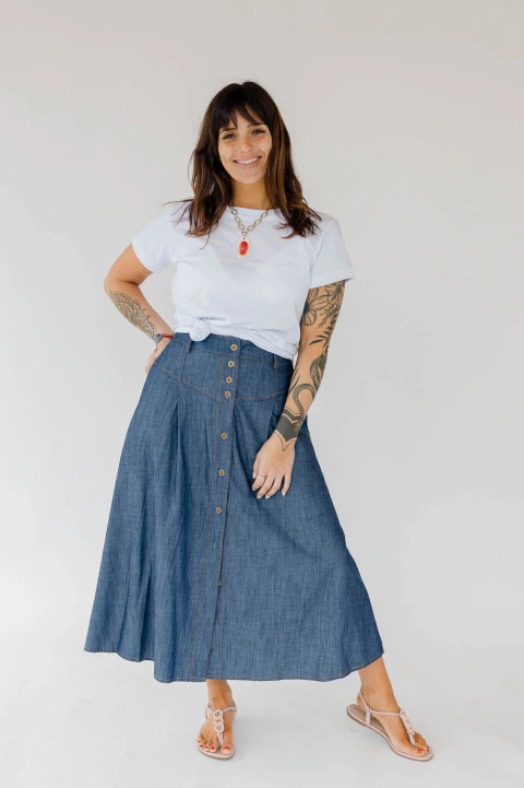 Mujer luciendo una pollera larga de denim azul con botones frontales, combinada con remera blanca y sandalias, sobre fondo claro.