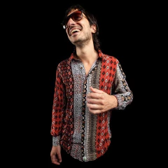 Hombre sonriente con camisa de manga larga estampada en tonos rojos, blancos y marrones, usando gafas de sol oscuras, fondo negro