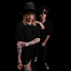 Dos modelos visten camisetas negras de manga corta, uno de ellos lleva un sombrero y gafas de sol, con un fondo negro.
