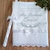Toalha de batizado personalizada em branco com bordado cinza, incluindo o nome "Enrico" e detalhes em fitas e rendas.