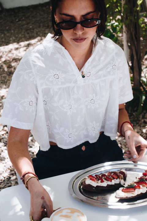 Mujer usando blusa corta Sicilia de poplin bordado blanca con volados, sentada en una mesa al aire libre con café y tostadas.