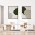 Dois quadros decorativos com formas abstratas em tons de verde e bege, pendurados em uma parede branca, ao lado de uma mesa de madeira clara e cadeiras minimalistas.