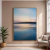 Quadro decorativo em canvas com imagem abstrata do mar azul ao pôr do sol, pendurado em parede bege ao lado de planta e poltrona