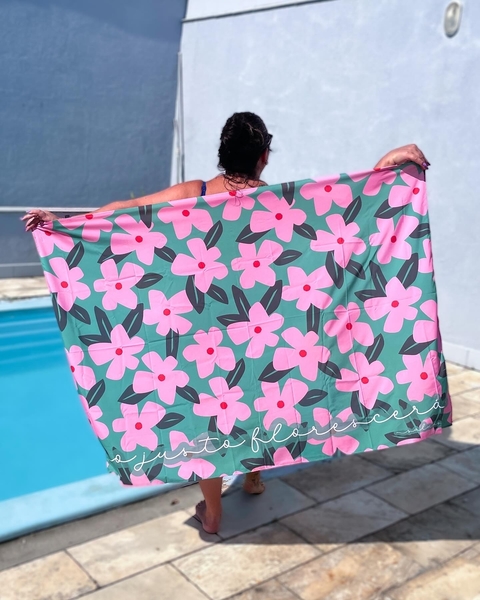 Canga de praia personalizada com estampa de flores rosas sobre fundo verde sendo segurada por mulher ao lado de piscina