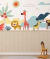Painel de parede infantil colorido com ilustrações de animais do safari, incluindo leão, girafa, zebra e macaco, decorando um quarto com parede bege e brinquedo de cavalo de madeira.