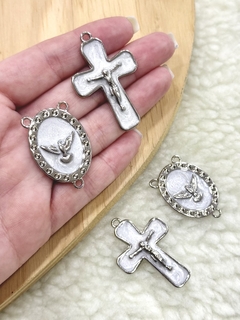 Quatro peças de entremeio em níquel perolado, incluindo dois crucifixos e dois medalhões com detalhes religiosos, seguradas por uma mão.