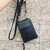 Mini cartera negra Avila colgando con correa en un fondo de pared de piedra.