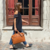 Mujer caminando por una acera de adoquines frente a un edificio con una puerta de madera y cortinas rojas, llevando un maletín de cuero marrón Udine. 
