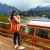 Mujer de espaldas con una tote bag color suela, disfrutando de una vista panorámica de montañas y un lago en un día nublado.