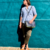 Una mujer apoyada contra una pared turquesa, luciendo una bandolera azul combinada y una camisa de manga larga.