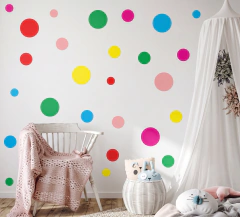 Adesivo de parede infantil com bolinhas coloridas em tons de vermelho, verde, azul, amarelo, rosa e rosa escuro em parede branca de quarto decorado com cadeira branca e manta rosa.