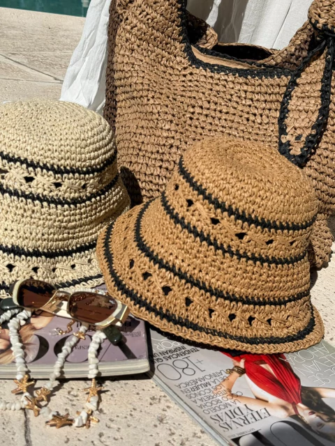 Sombreros de rafia modelo Caipi en tonos beige y marrón con líneas negras, junto a una bolsa tejida del mismo material, lentes de sol y revistas, sobre una superficie al aire libre.