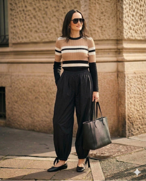 Pantalón tipo babucha negro de textura arrugada, con cintura elástica y lazos en los tobillos, combinado con suéter de rayas y cartera negra. Mujer de pie en una calle urbana.