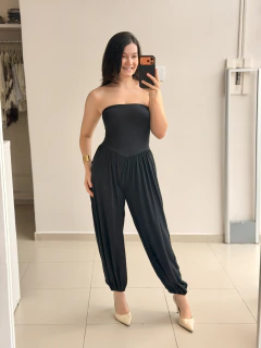 Mulher sorridente vestindo macacão preto tomara que caia com pernas largas estilo Aladim, posando em ambiente interno claro com piso de cerâmica.