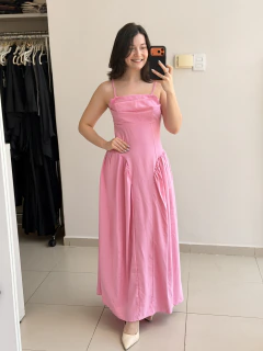 Mulher sorridente vestindo vestido longo rosa com alças finas e decote reto, posando em frente a um espelho em ambiente interno