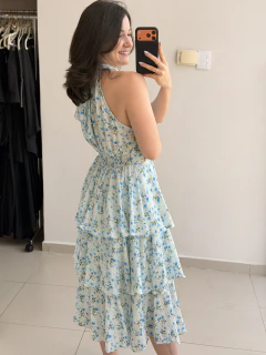 Mulher sorridente usando vestido babado com estampa de pequenas rosas azuis em fundo claro, tirando selfie de costas em ambiente interno