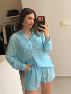 Mulher usando conjunto azul claro Uiara Short, composto por camisa over de viscose leve e short com lastex, tirando selfie em ambiente interno.
