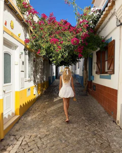 Mulher de costas vestindo vestido branco curto Nyx, caminhando por rua de pedra com flores rosa acima